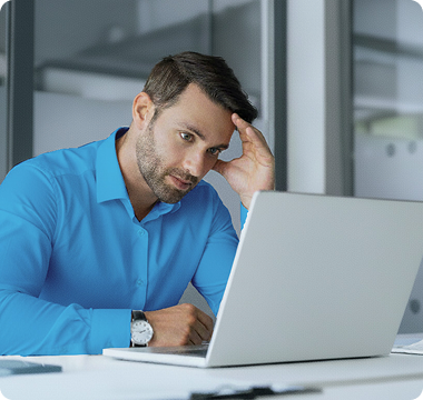 A man in a blue shirt looks at a laptop with a concerned expression, resting his head on one hand. The office setting is modern and minimalistic.