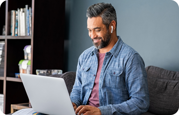 Smiling man in a blue shirt sitting in a living room and working on a laptop with earbuds in
