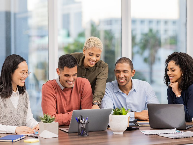 Team of five smiling coworkers gathered around a laptop, planning together at a shared desk