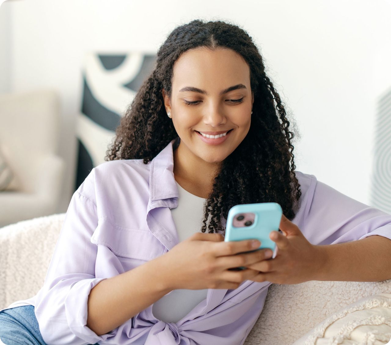 A woman with curly hair, wearing a lavender shirt, smiles while looking at her phone. She's seated on a beige couch, creating a relaxed atmosphere.
