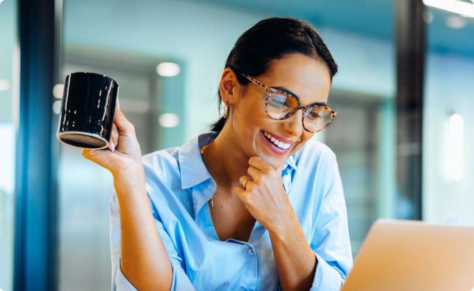 A woman in glasses and a blue shirt smiles while holding a black coffee mug, looking at a laptop. She appears cheerful and engaged in a bright office.