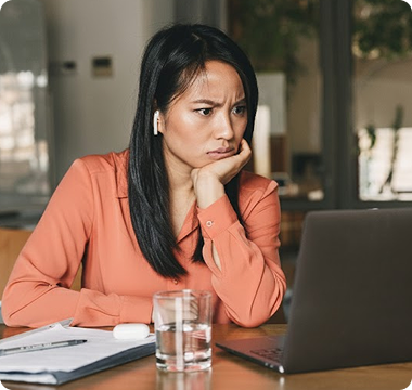 A woman in an orange blouse, looking frustrated or deep in thought, sits at a table with a laptop, glass of water, and papers in a bright office space.