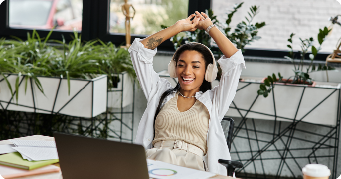Young woman sitting at a desk, wearing headphones, and stretching with a joyful expression. Relaxed office setting with plants around her.