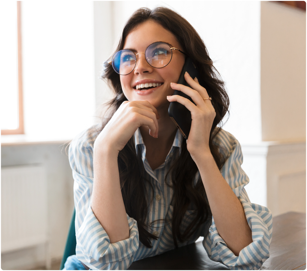 Young woman in glasses and a striped shirt smiles while talking on the phone, seated at a wooden table in a bright, airy room. Mood is cheerful.