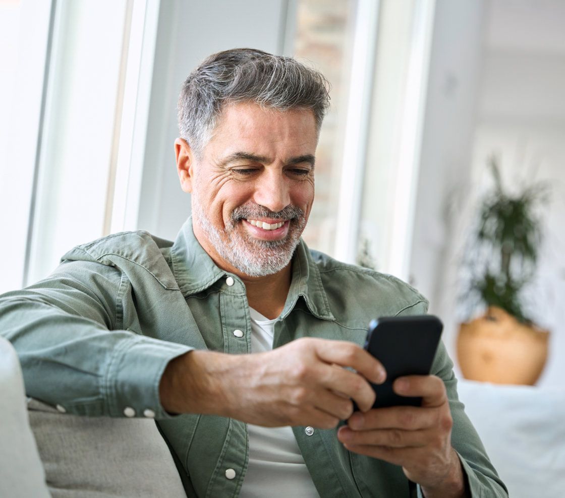Middle-aged man with grey hair and a beard smiling at a smartphone. He's relaxed on a sofa, wearing a green shirt. The room is bright with a plant in the background.