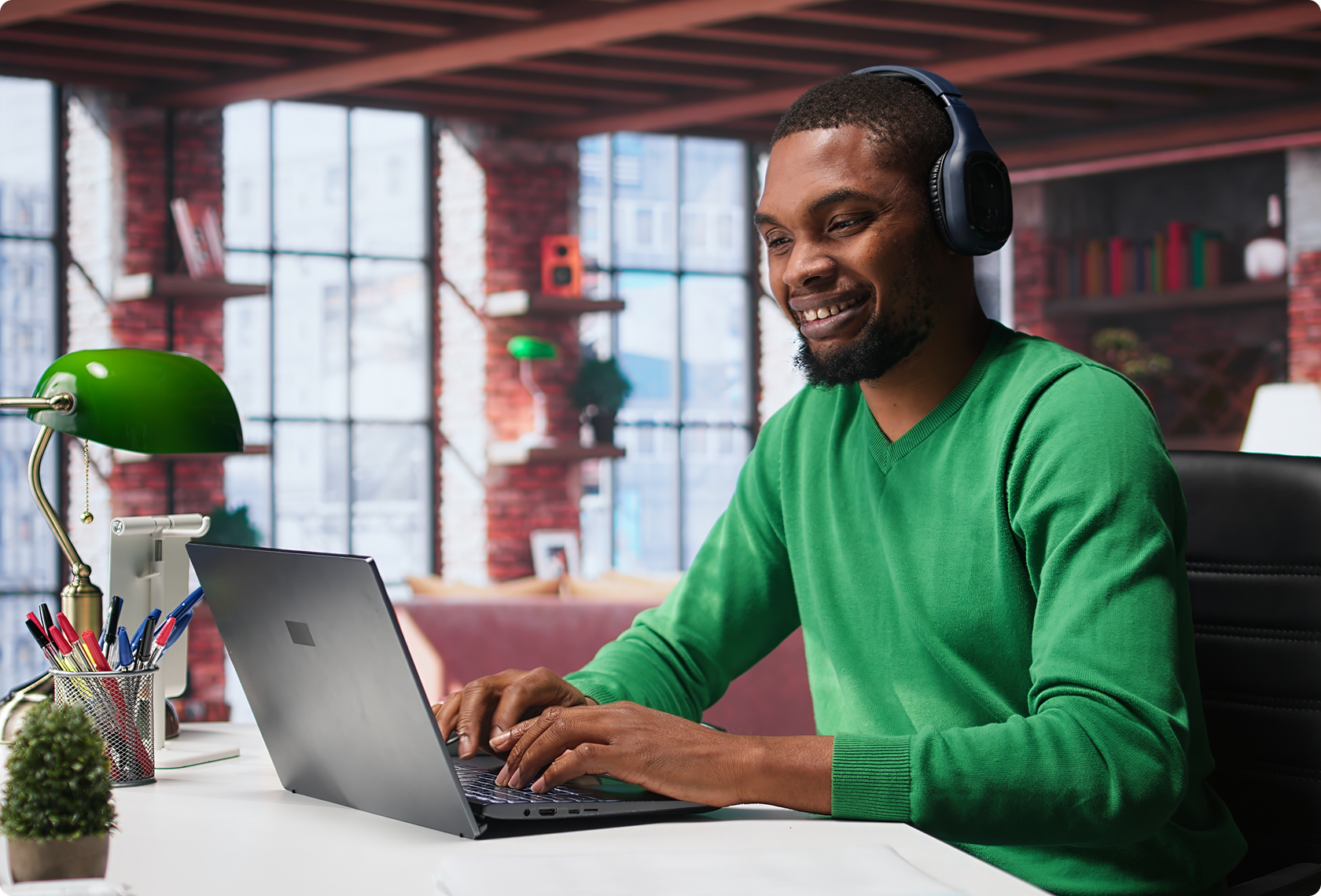 Smiling man in green sweater works on laptop with headphones in a stylish room with exposed brick and large windows, conveying a productive vibe.