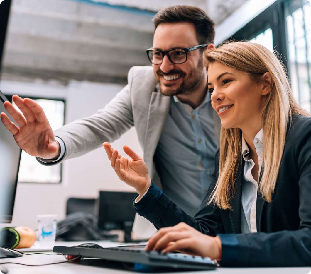 A man and a woman in business attire collaborate at a computer, smiling and gesturing at the screen. The scene is bright and conveys teamwork.
