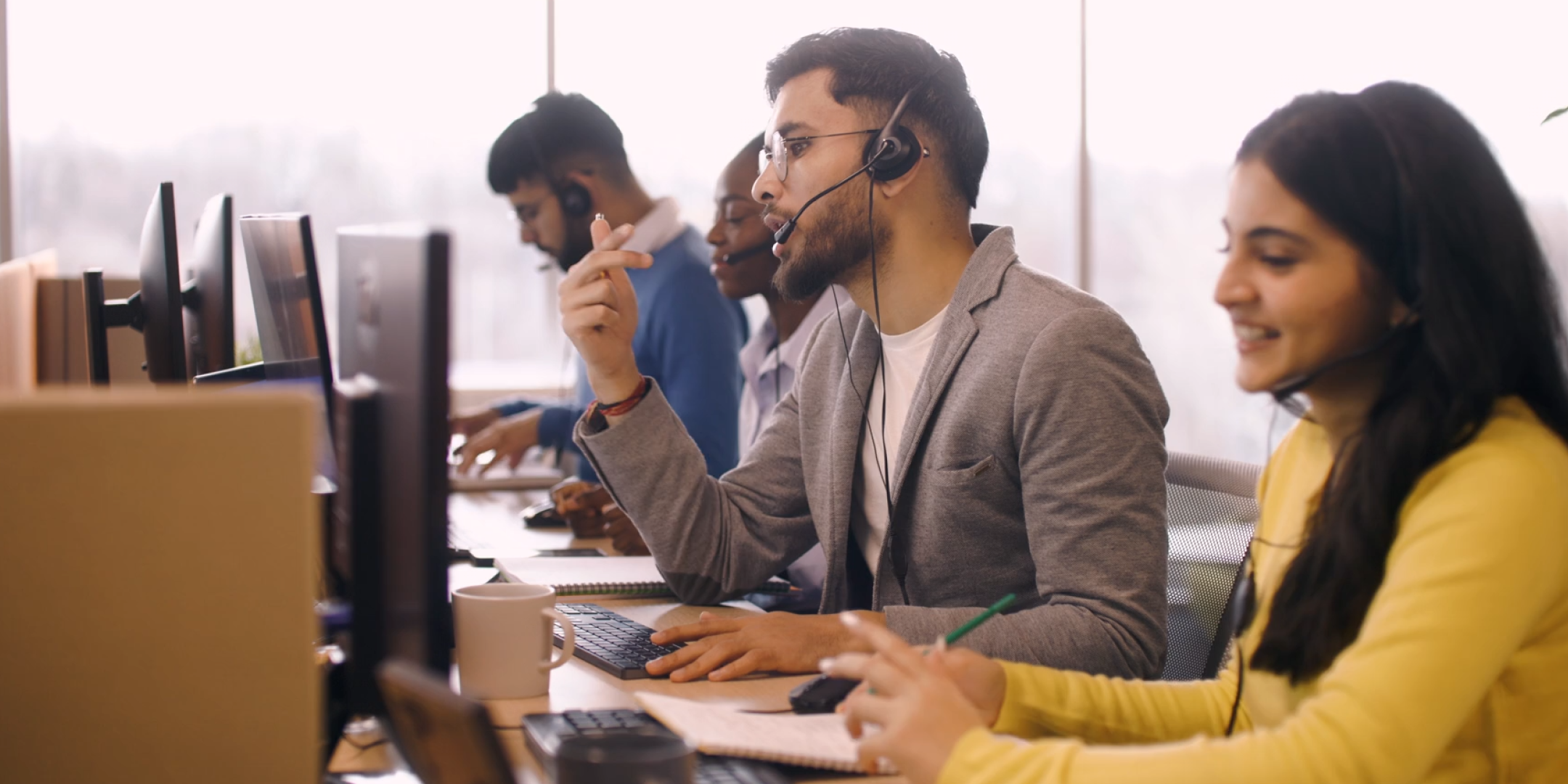 A diverse group of people work at computers in an office, wearing headsets. They appear engaged, suggesting a collaborative and focused environment.