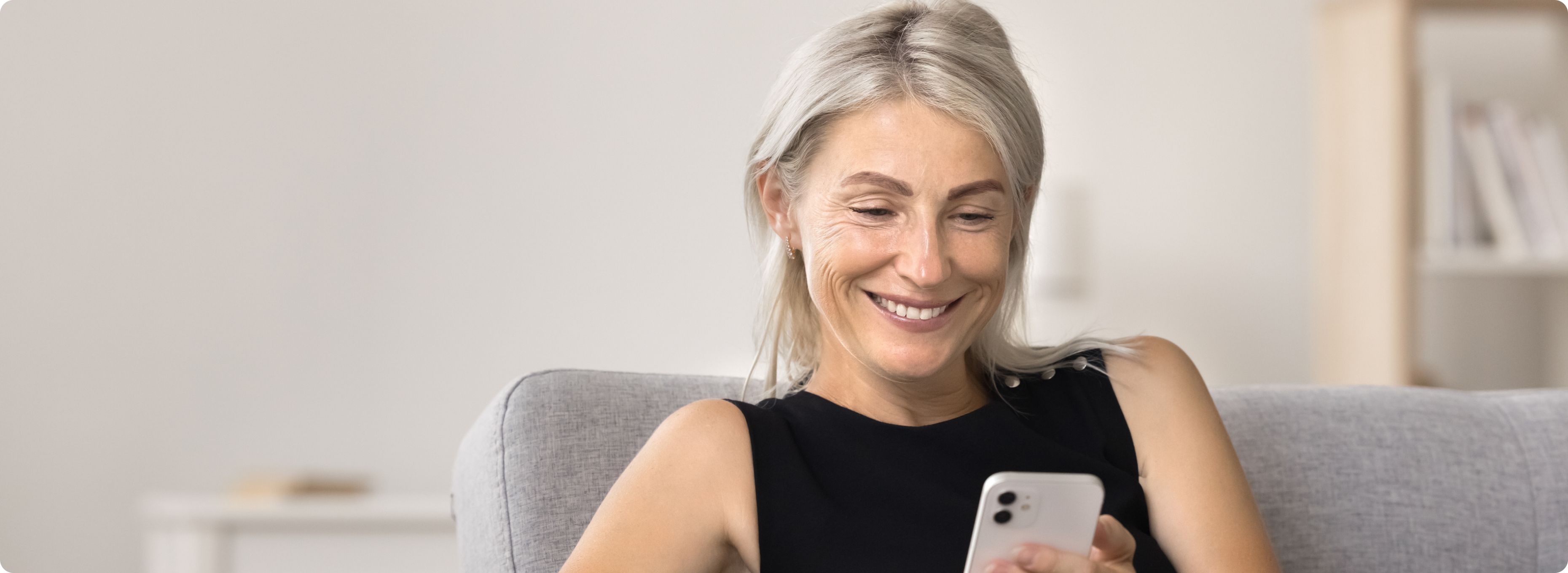 A woman with gray hair smiles while sitting on a couch, looking at her smartphone. She wears a black sleeveless top in a bright, cozy room.