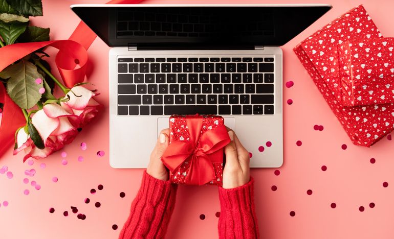Someone holding a valentine's day gift over a laptop on a desk scattered with valentine's day decorations