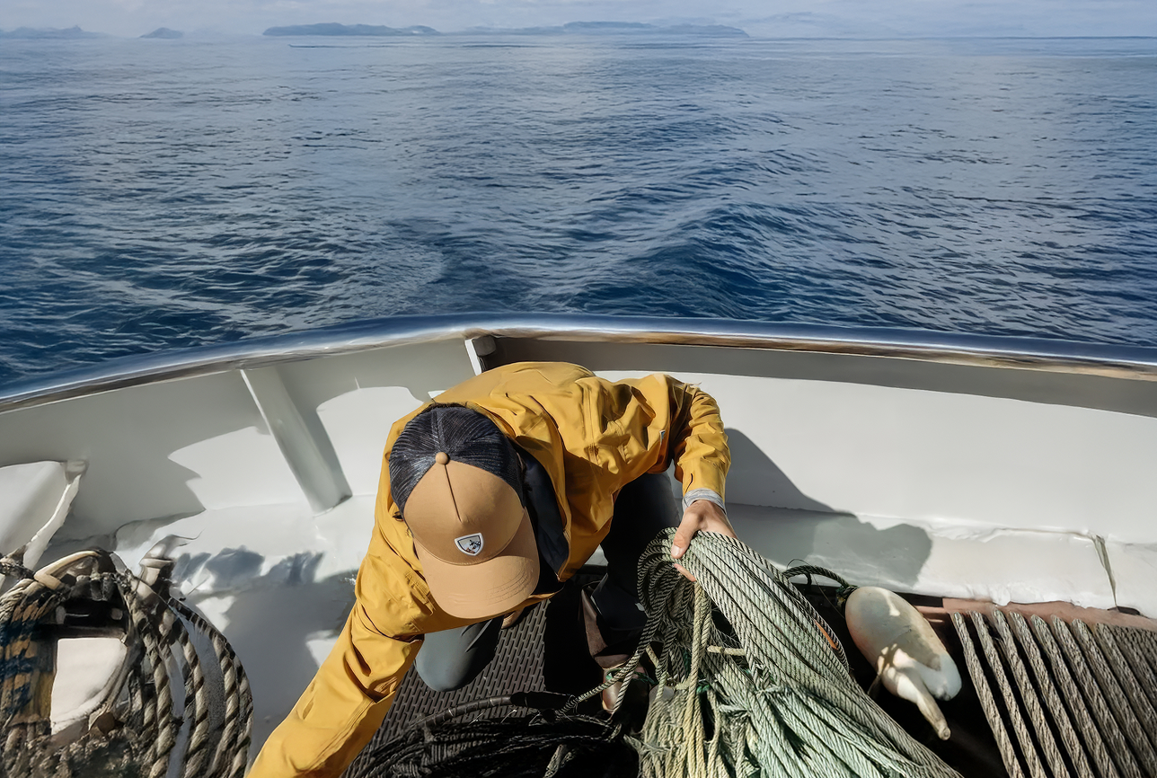 A person in a yellow jacket and cap organizes ropes on a boat deck against a backdrop of calm ocean and distant islands, conveying a sense of adventure.