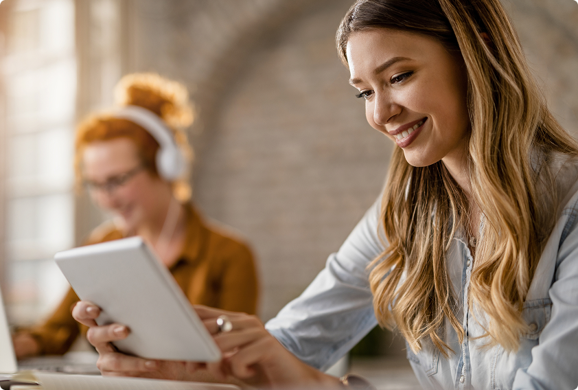 A woman in a light blue shirt smiles while using a tablet in a bright, casual office. In the background, another woman with headphones focuses on a laptop.