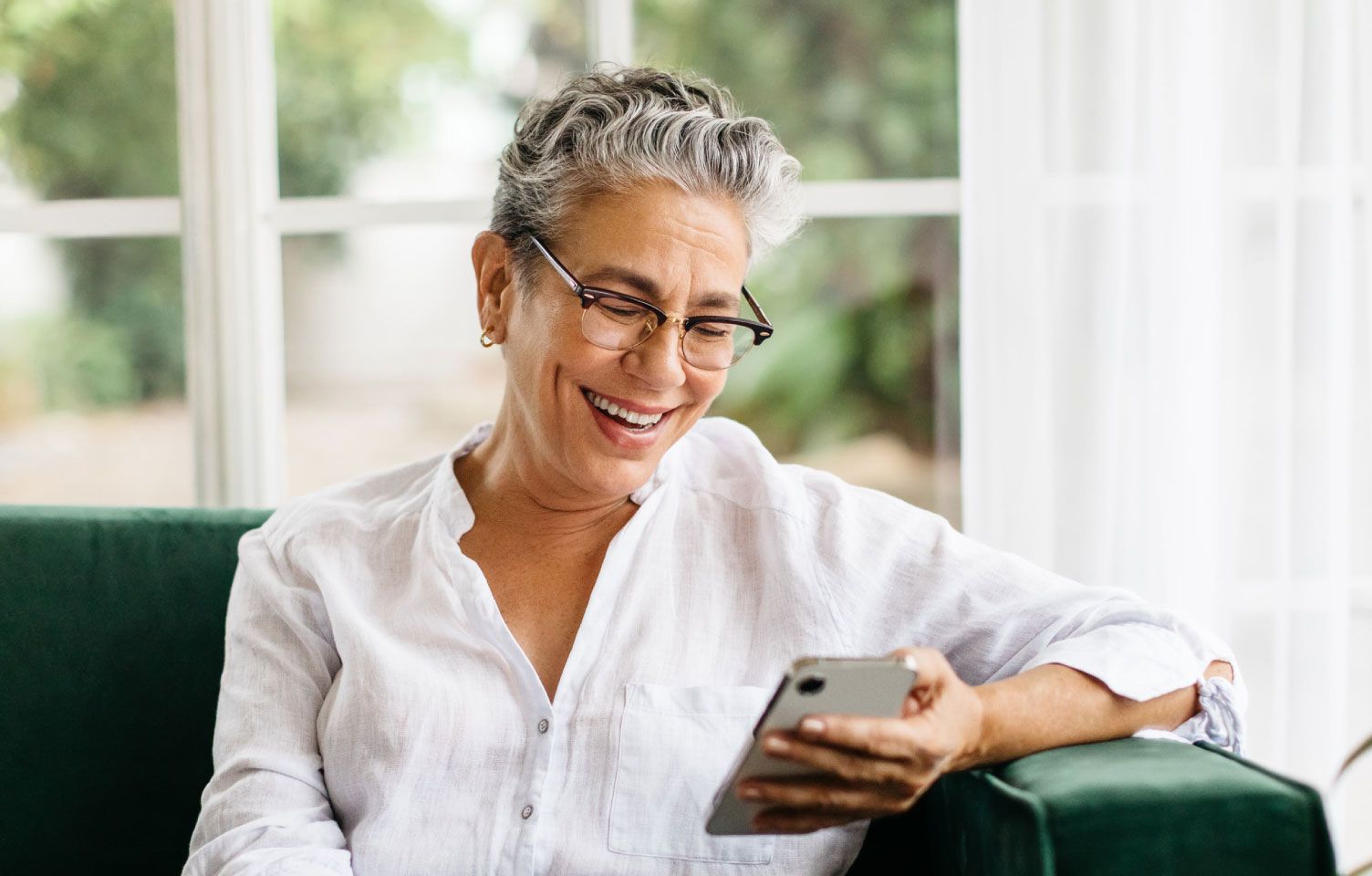 Elderly person with short gray hair and glasses, smiling while looking at a smartphone. Seated on a green couch with a bright window in the background.