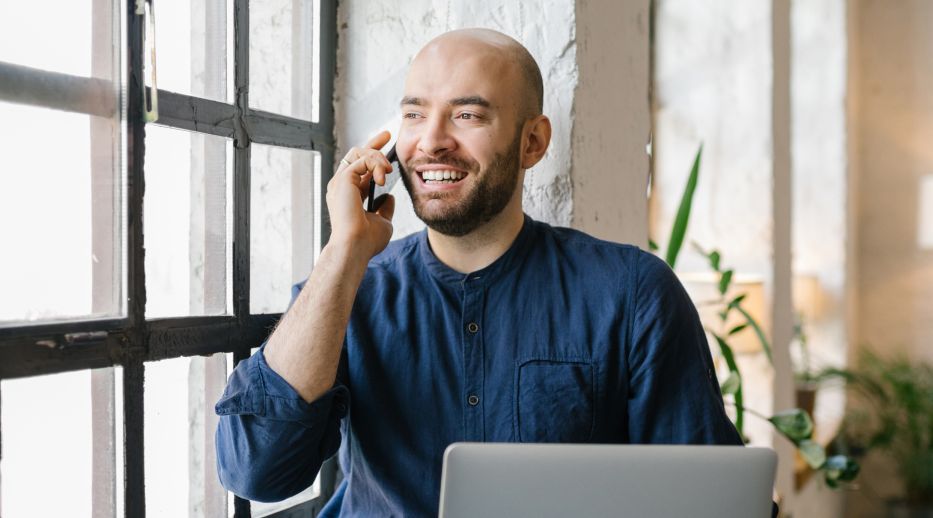 A smiling man in a blue shirt talks on a phone near a large window, with a laptop on the table. The setting is bright, cozy, and relaxed.