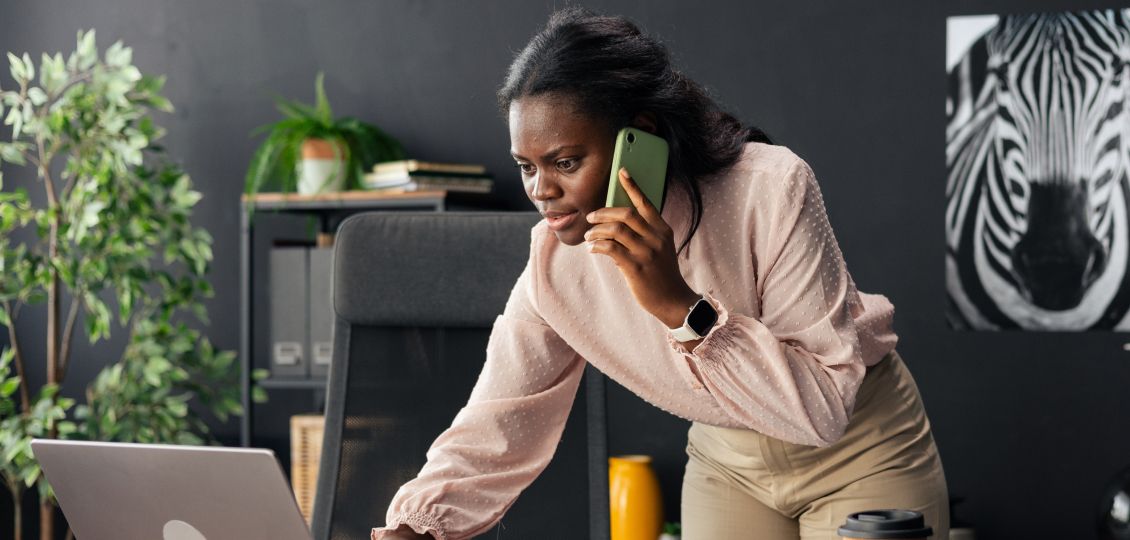 Woman in an office leans over a laptop while talking on a phone. She's wearing a smartwatch, and there's a zebra poster and plants in the background.