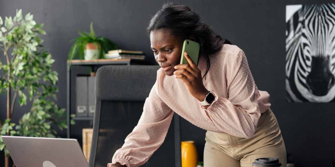 Woman in an office leans over a laptop while talking on a phone. She's wearing a smartwatch, and there's a zebra poster and plants in the background.