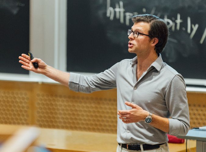 A man in glasses and a gray shirt is passionately lecturing in a classroom, gesturing with his hand. A chalkboard and link are visible behind him.