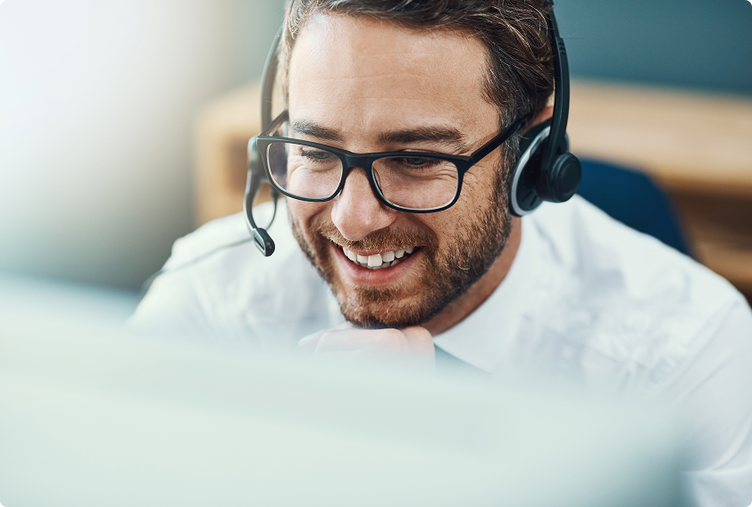 Smiling man with glasses and headset works at a computer in an office, conveying a friendly and professional customer service environment.
