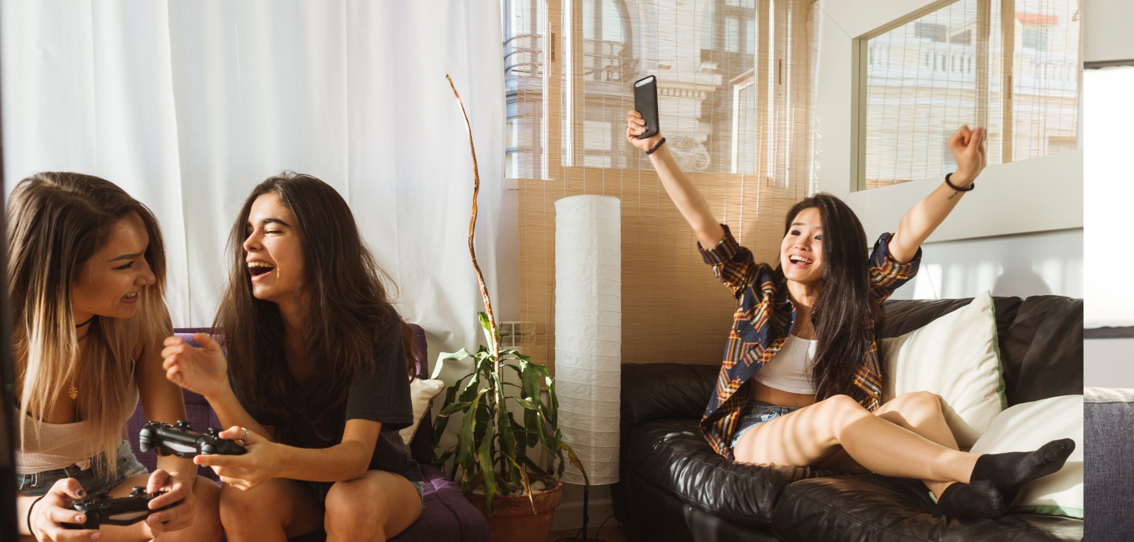 Three women are having fun in a bright living room. Two are playing video games and laughing, while the third cheers joyfully on a couch. Relaxed and lively mood.