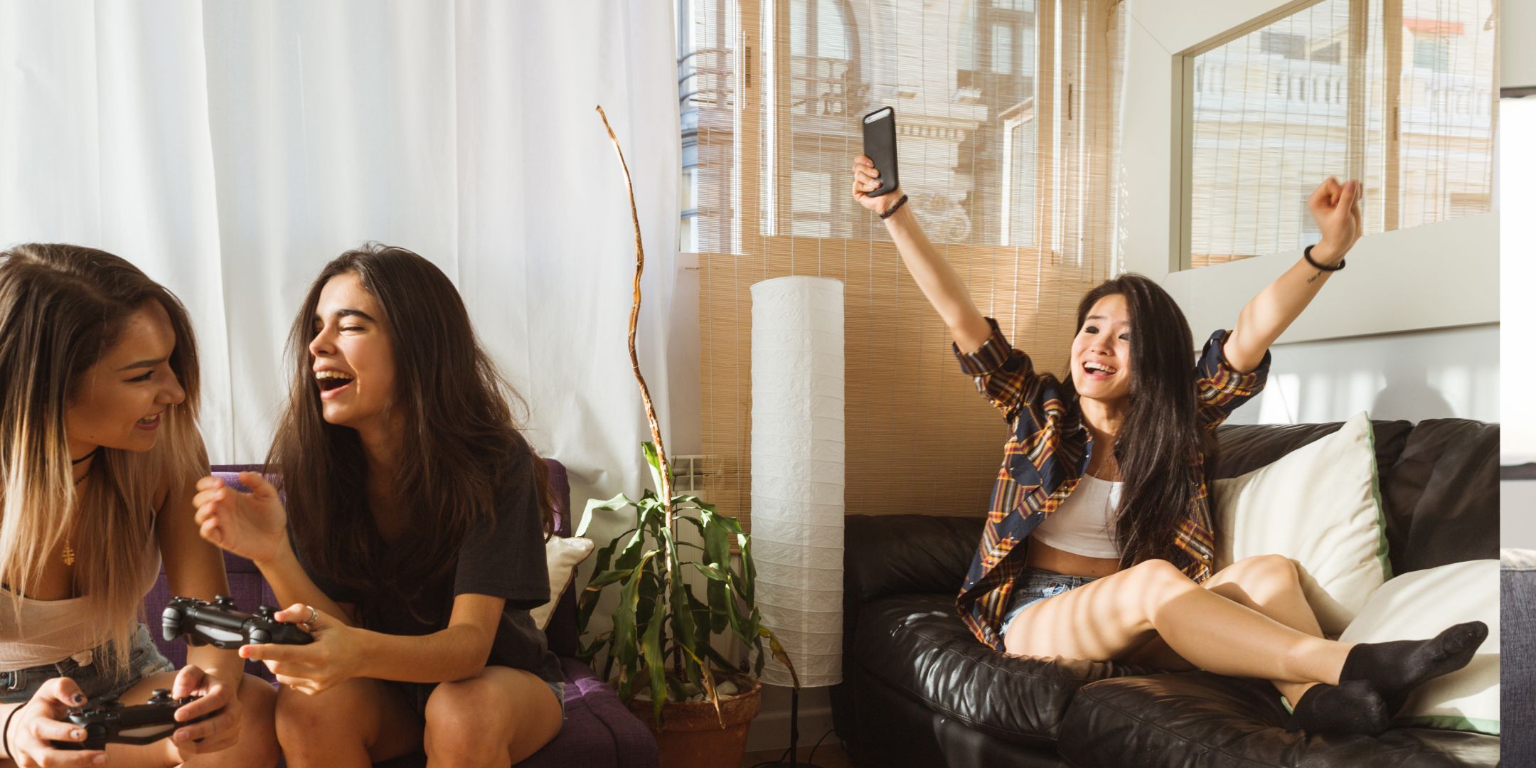 Three women are having fun in a bright living room. Two are playing video games and laughing, while the third cheers joyfully on a couch. Relaxed and lively mood.