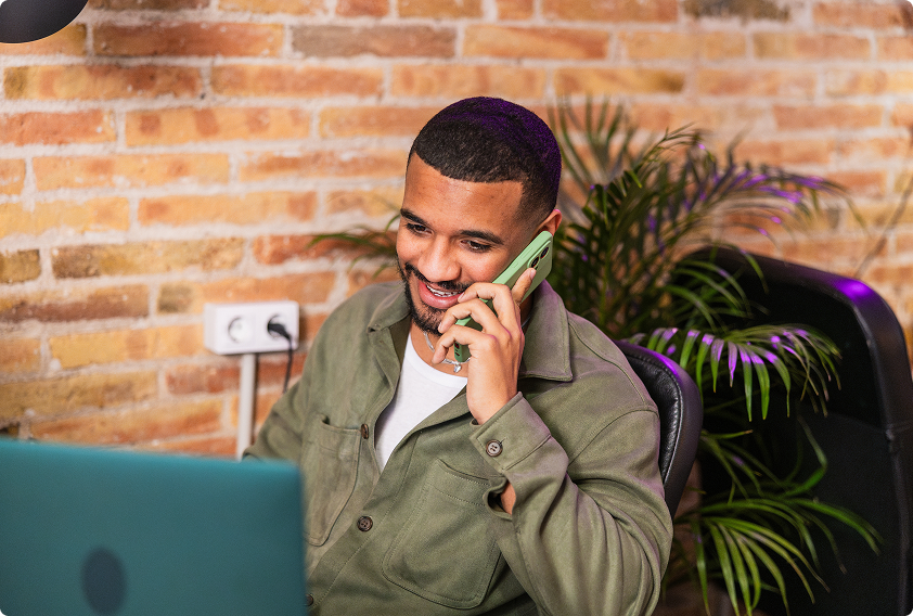 A man in a green jacket, smiling as he talks on a phone, sits at a desk with a laptop. A brick wall and potted plant are in the background.