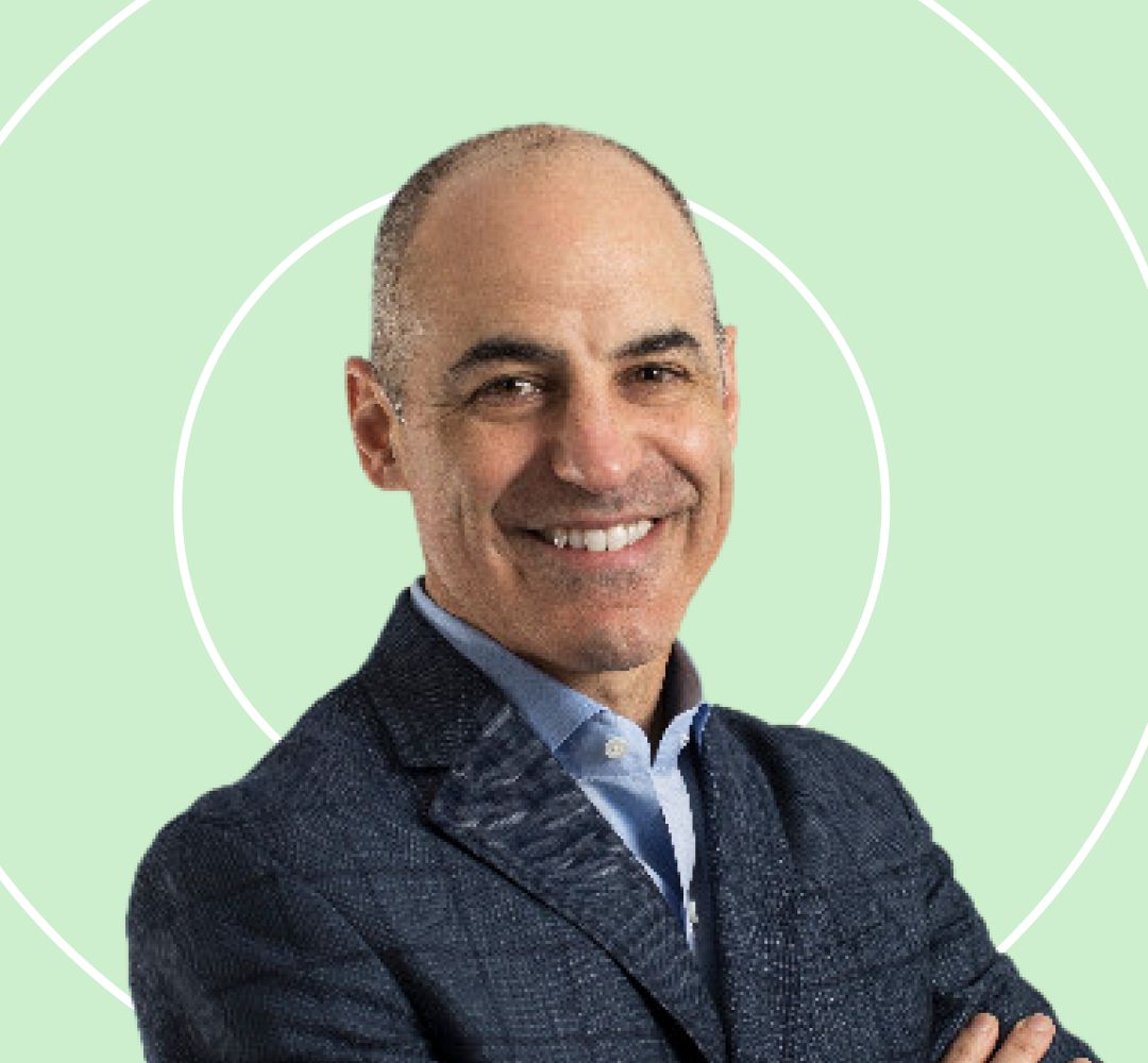 A man in a dark suit smiles confidently, standing with arms crossed against a light green background with subtle circular patterns.