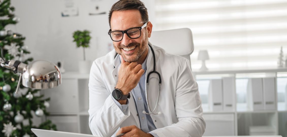 Smiling male doctor with glasses and stethoscope looks at a laptop in a bright, festive office with a decorated Christmas tree in the background.