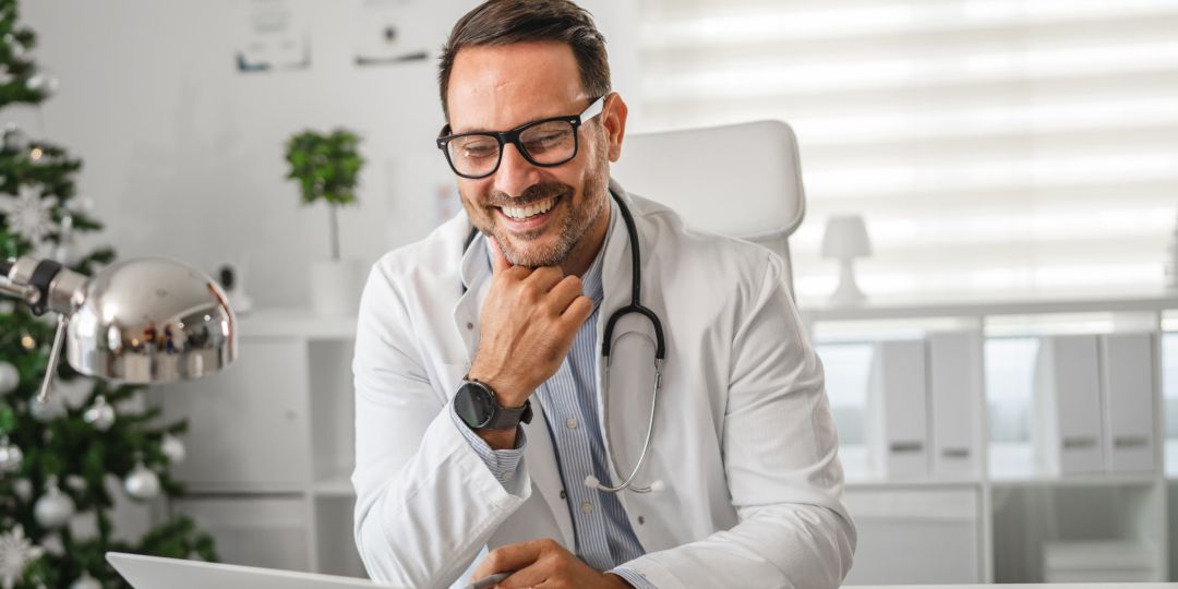 Smiling male doctor with glasses and stethoscope looks at a laptop in a bright, festive office with a decorated Christmas tree in the background.