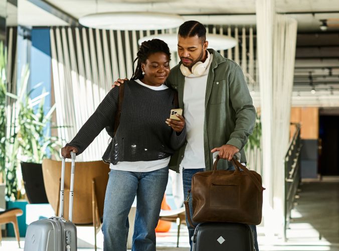A couple stands in a modern lobby, smiling at a phone. They have luggage, suggesting travel. The scene conveys a relaxed, happy atmosphere.