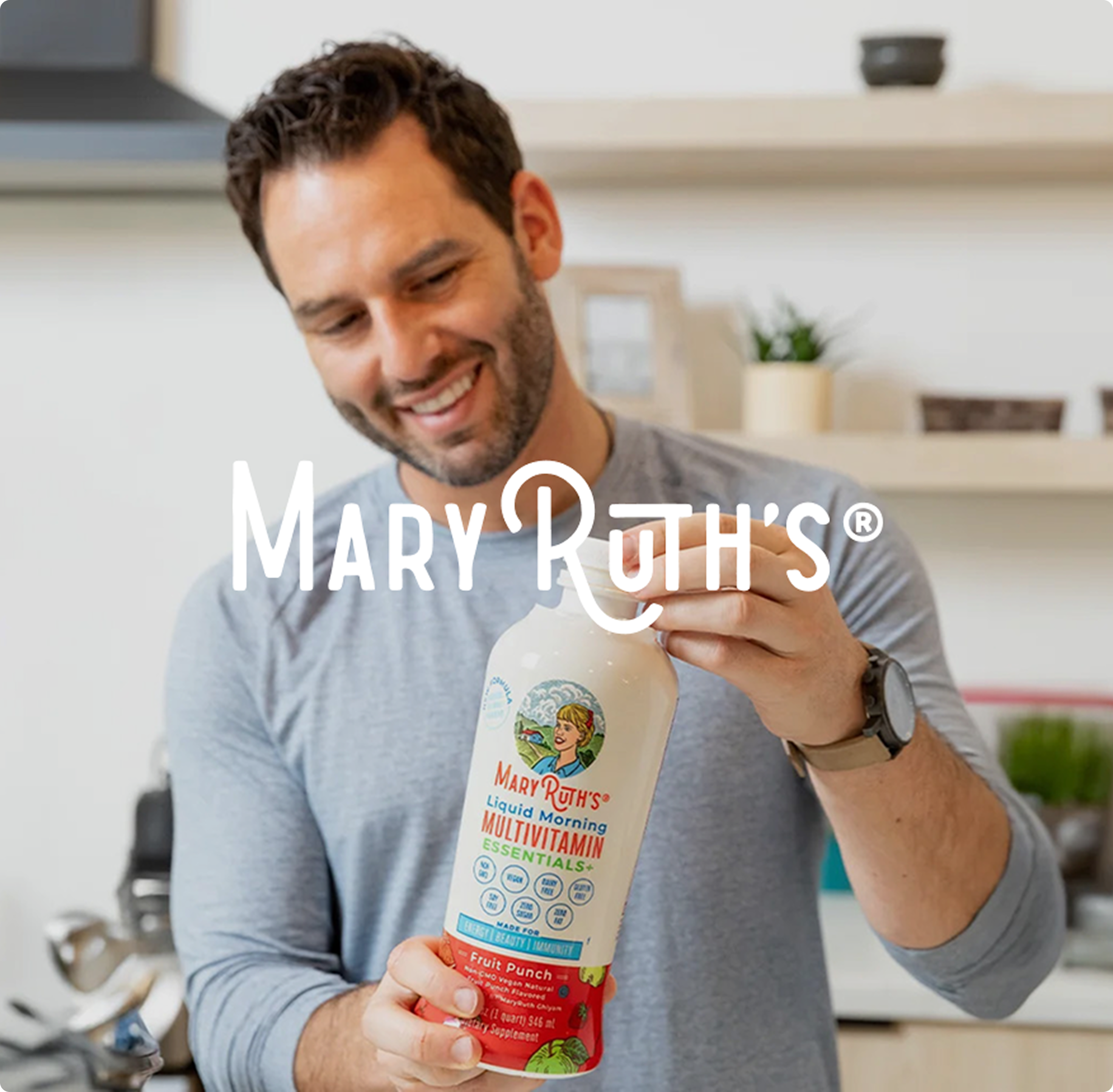 Smiling man in a kitchen holding a bottle of MaryRuth’s Liquid Morning Multivitamin. The atmosphere is bright and welcoming. Logo text overlaps.