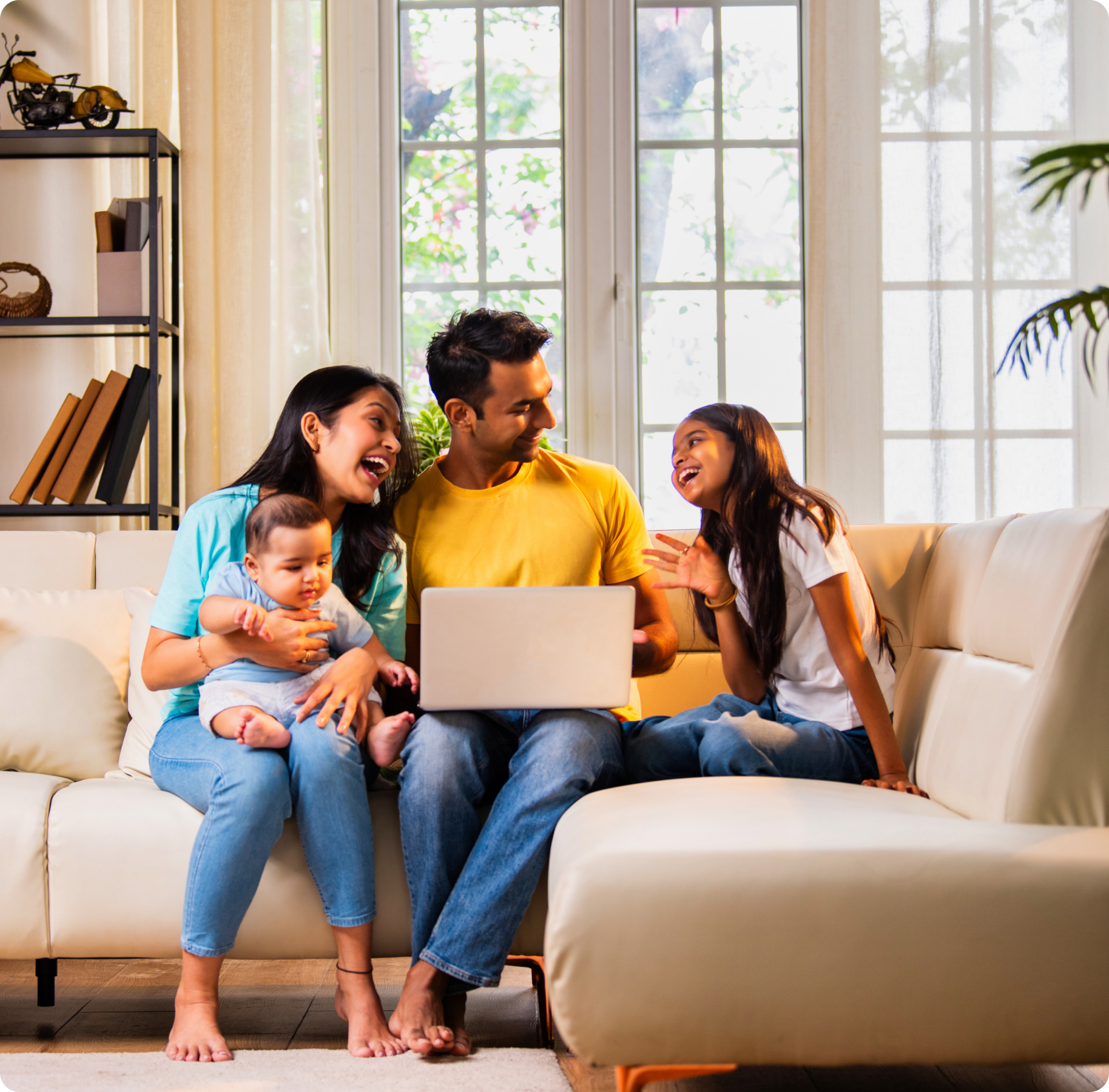 A joyful family of four sits on a beige sofa. Parents and two children gather around a laptop, smiling in a bright living room with large windows and plants.