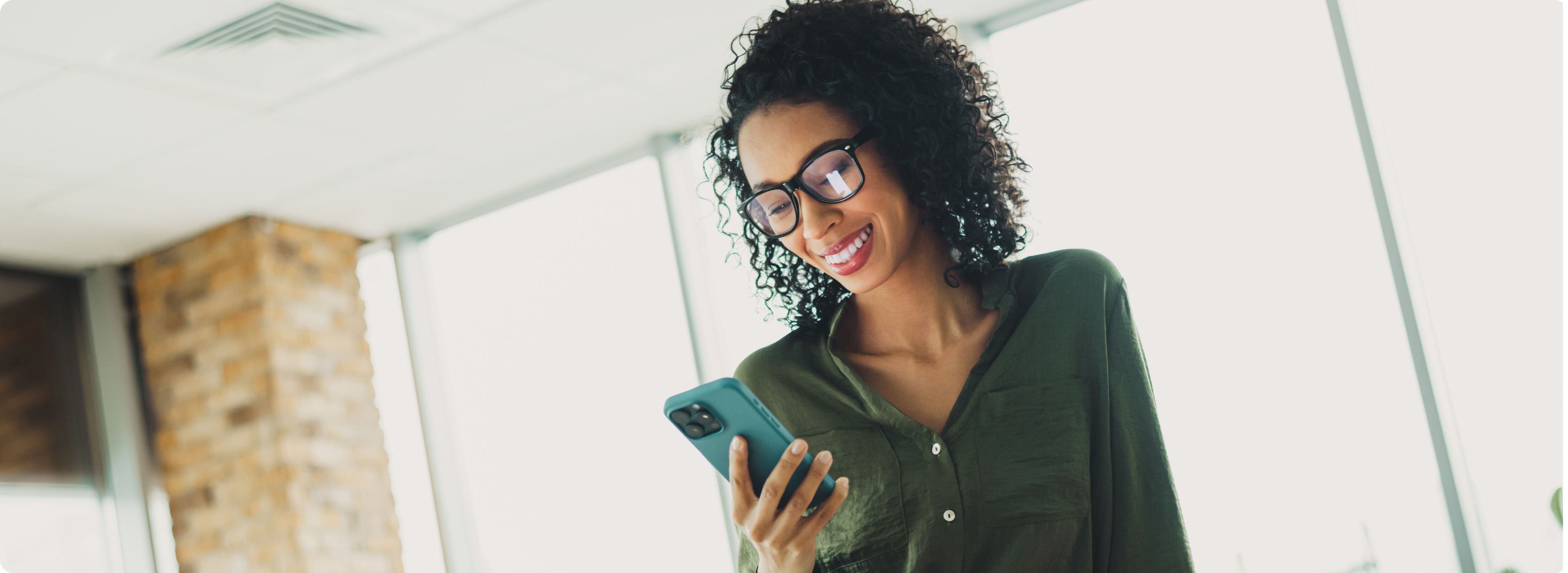 A woman with curly hair and glasses smiles while looking at her smartphone. She is wearing a green blouse, and the background is a bright office space.