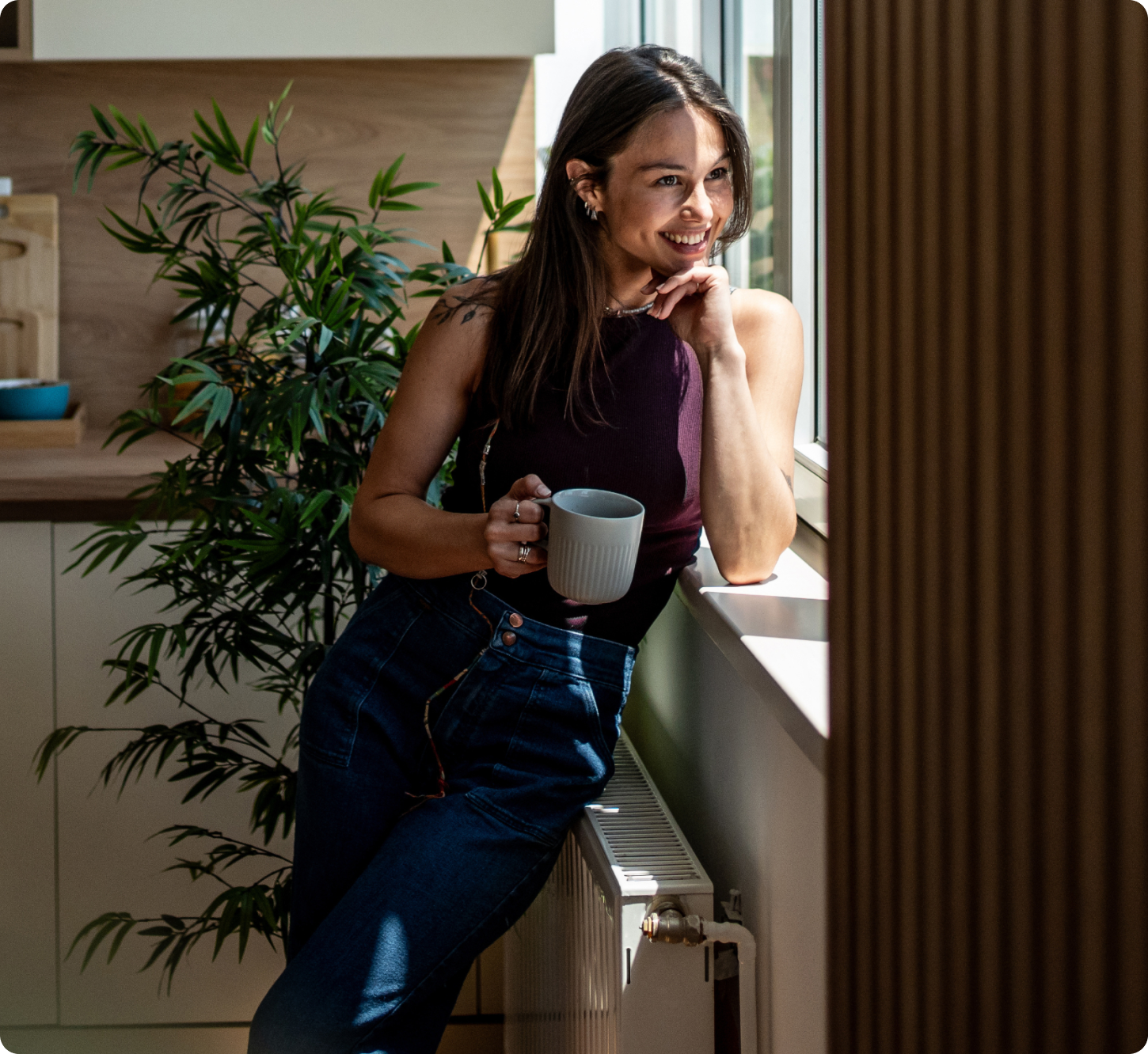 Smiling woman in a kitchen leaning on a windowsill, holding a mug. Sunlight filters in, illuminating her face. Warm, relaxed atmosphere.