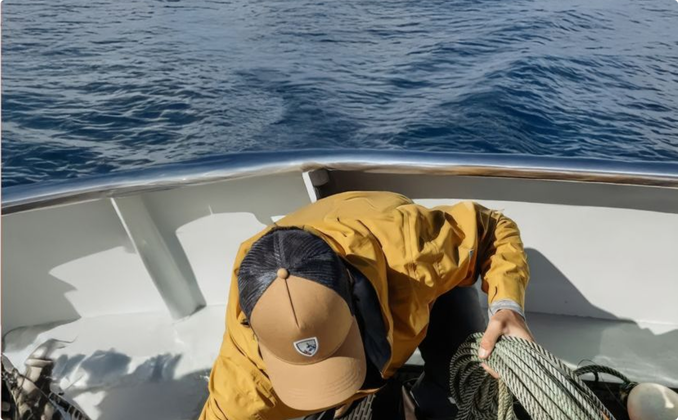 A person in a yellow jacket and cap organizes ropes on a boat deck against a backdrop of calm ocean and distant islands, conveying a sense of adventure.
