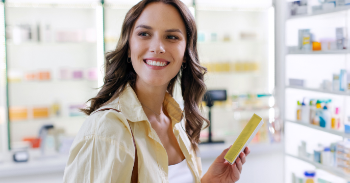 A smiling woman with wavy brown hair holds a yellow box inside a bright pharmacy. She wears a cream shirt, conveying a cheerful and welcoming atmosphere.