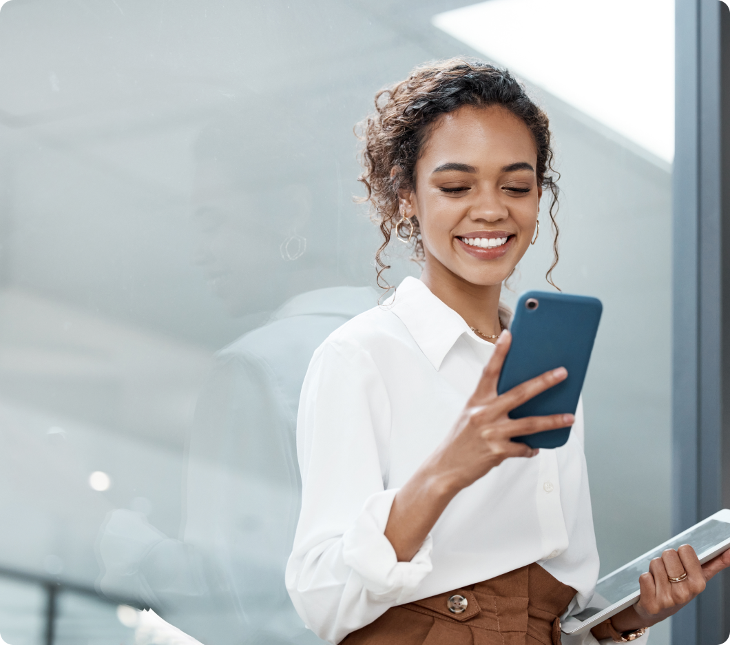 Smiling woman in a white blouse holds a phone, with her reflection in glass. She appears focused and cheerful in a modern indoor setting.