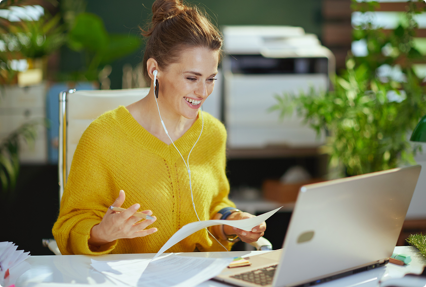 A woman in a yellow sweater smiles while on a video call at a desk. She wears earbuds and holds papers. The background is a bright, plant-filled office.
