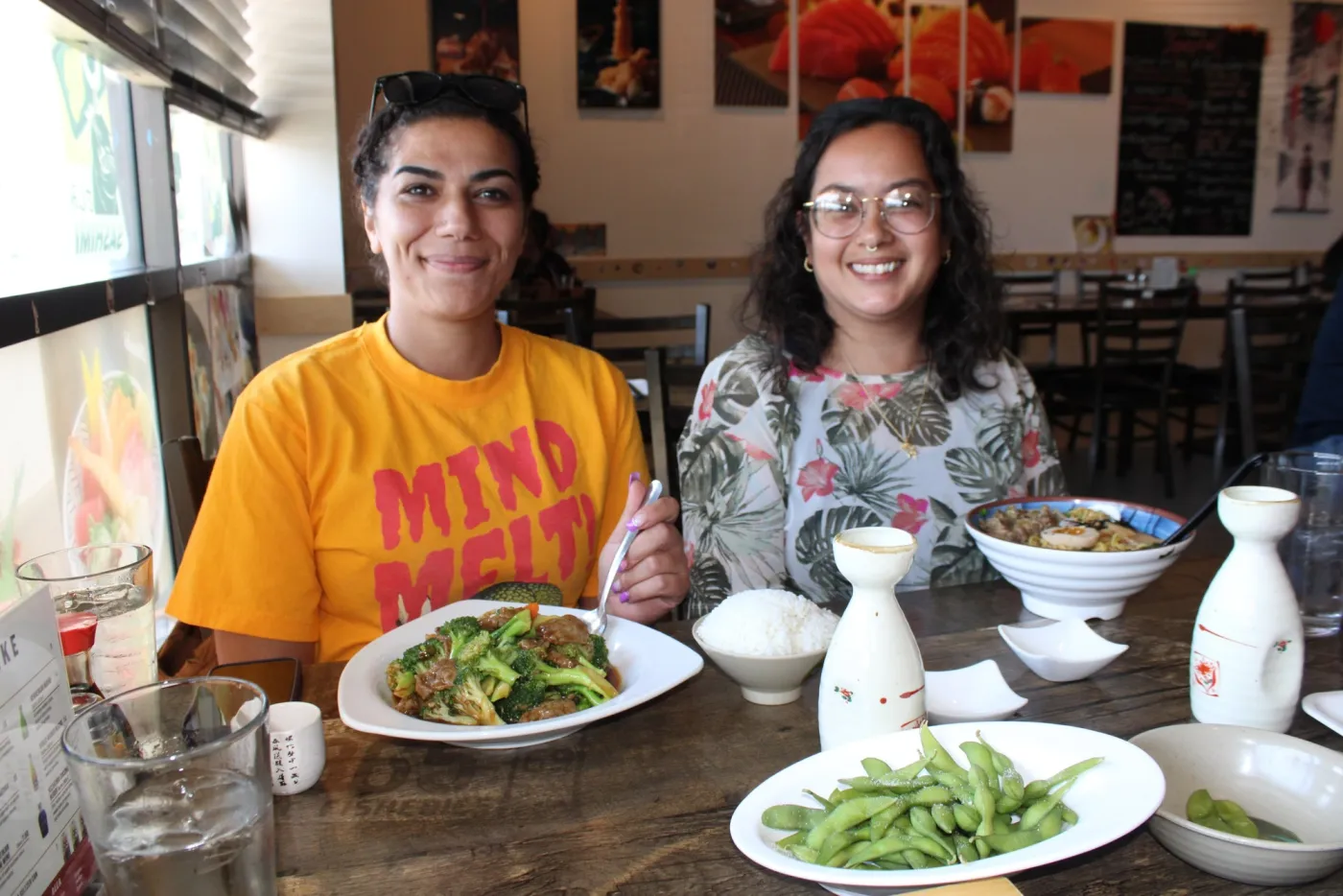 Two people smiling at a sushi restaurant table set with rice, beef and broccoli, edamame, ramen, and sake.