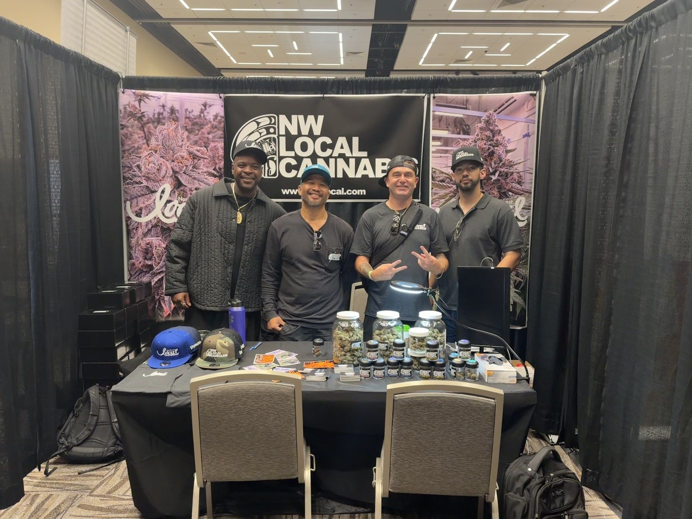 Four members of the Northwest Local Cannabis team standing behind their booth table of flower jars under the NW Local Cannabis banner.