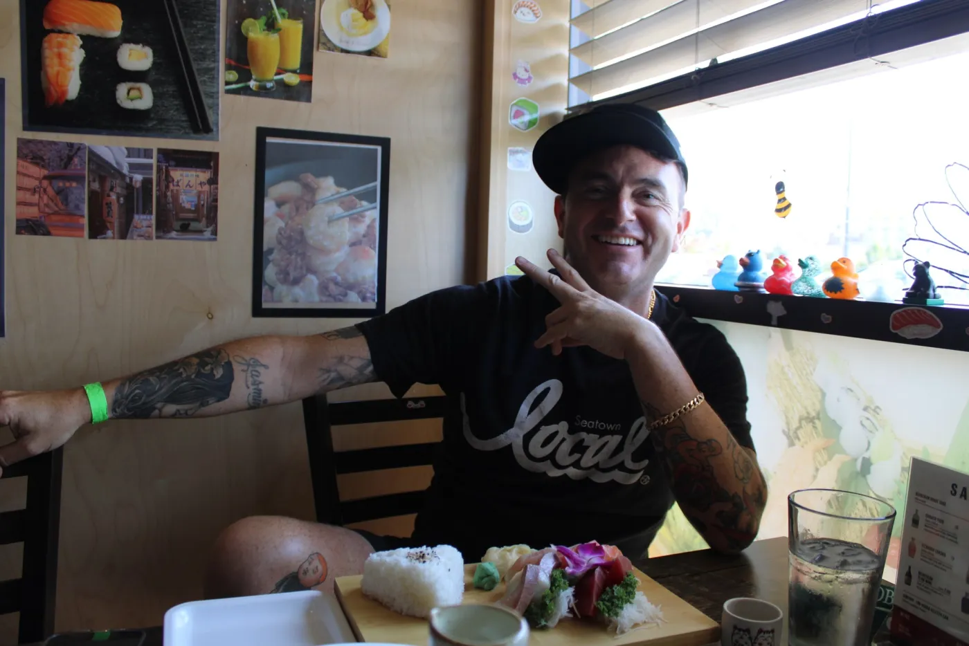 A person seated at a sushi restaurant giving a peace sign, with a board of sashimi and rice in front of them.