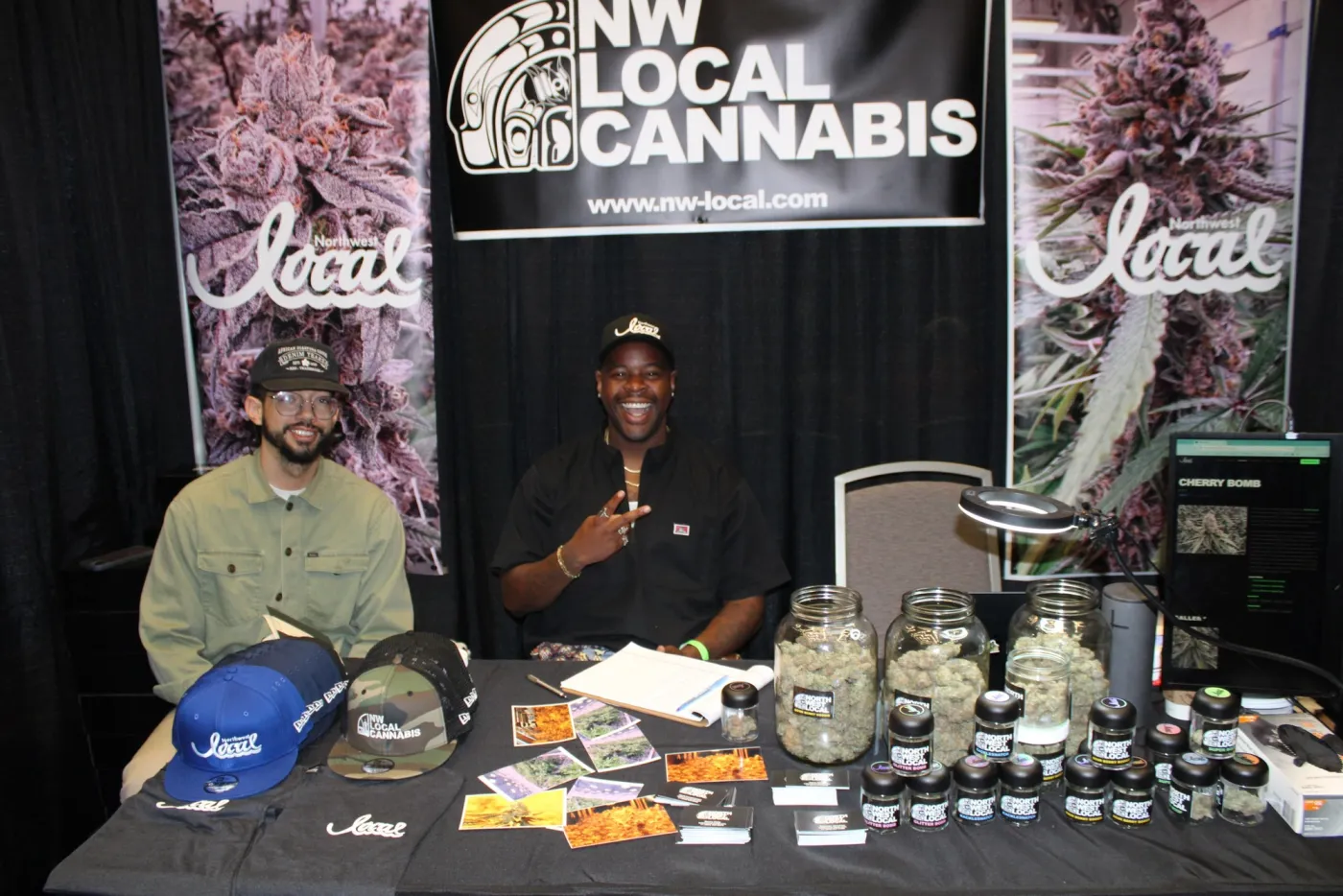 Two Northwest Local team members seated at the booth behind jars of flower, one giving a peace sign, under the banner.