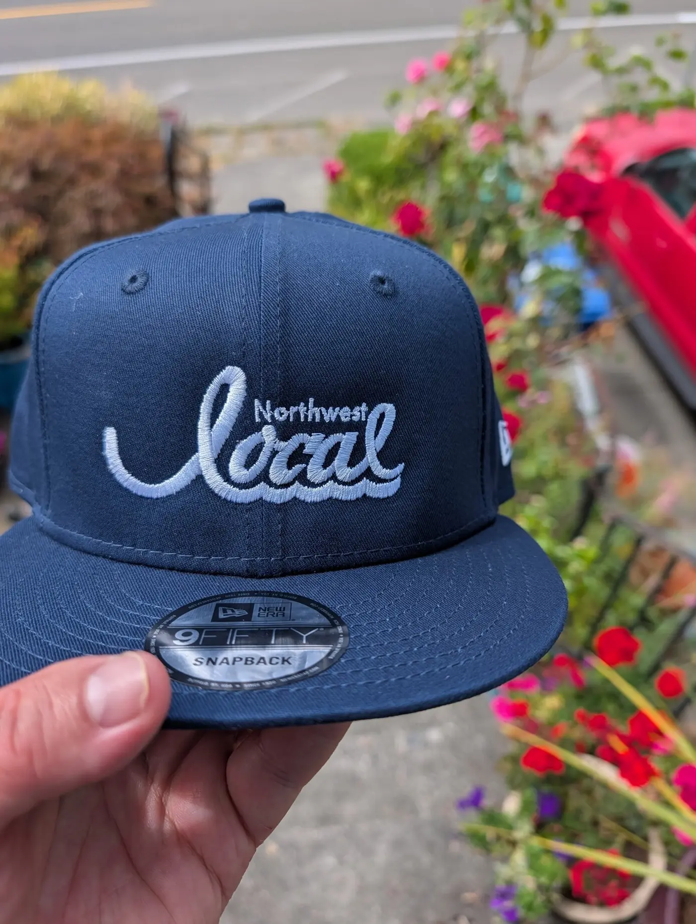 A navy New Era snapback with the Northwest Local script logo, held up outdoors in front of roses.