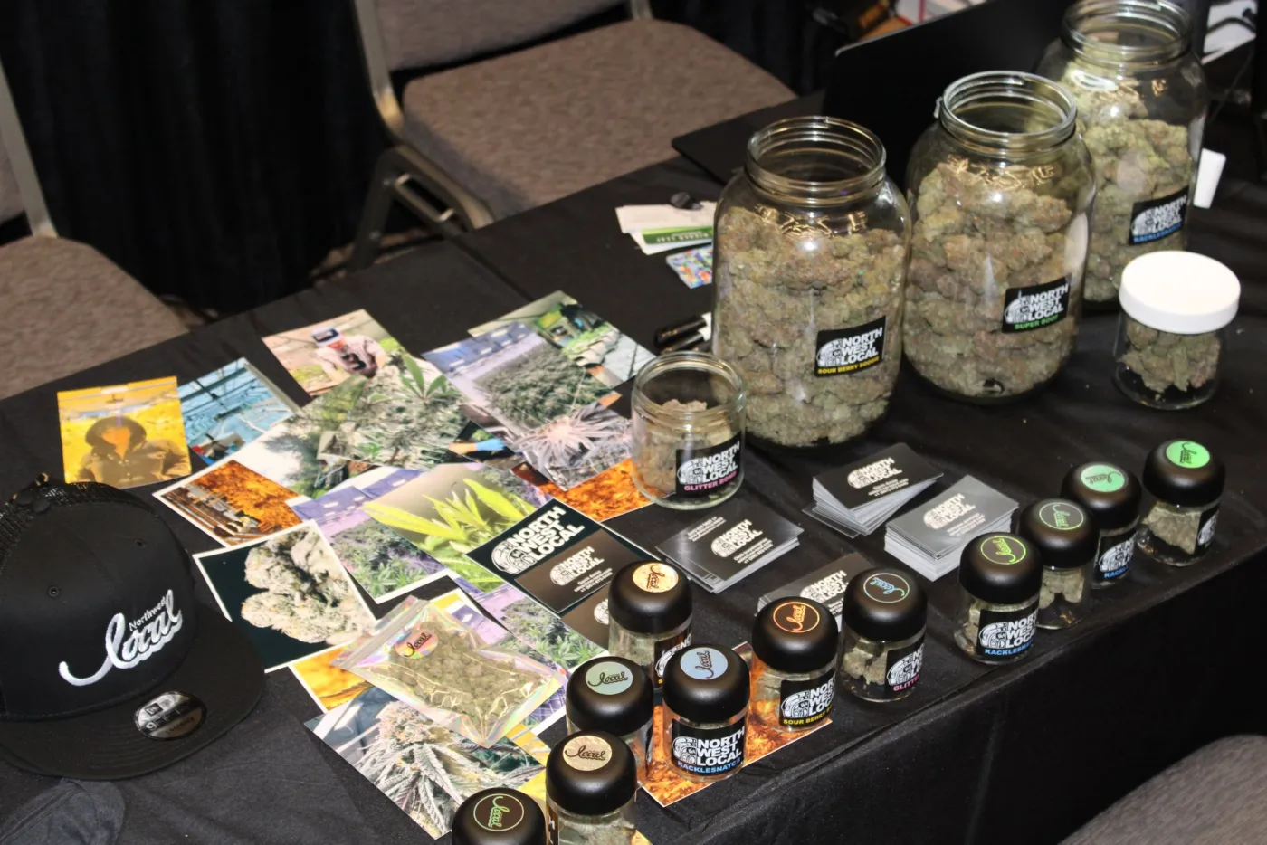 A booth table spread with large jars of flower labeled Sour Berry Boogie and Super Boof, smaller jars, printed photos, and business cards.