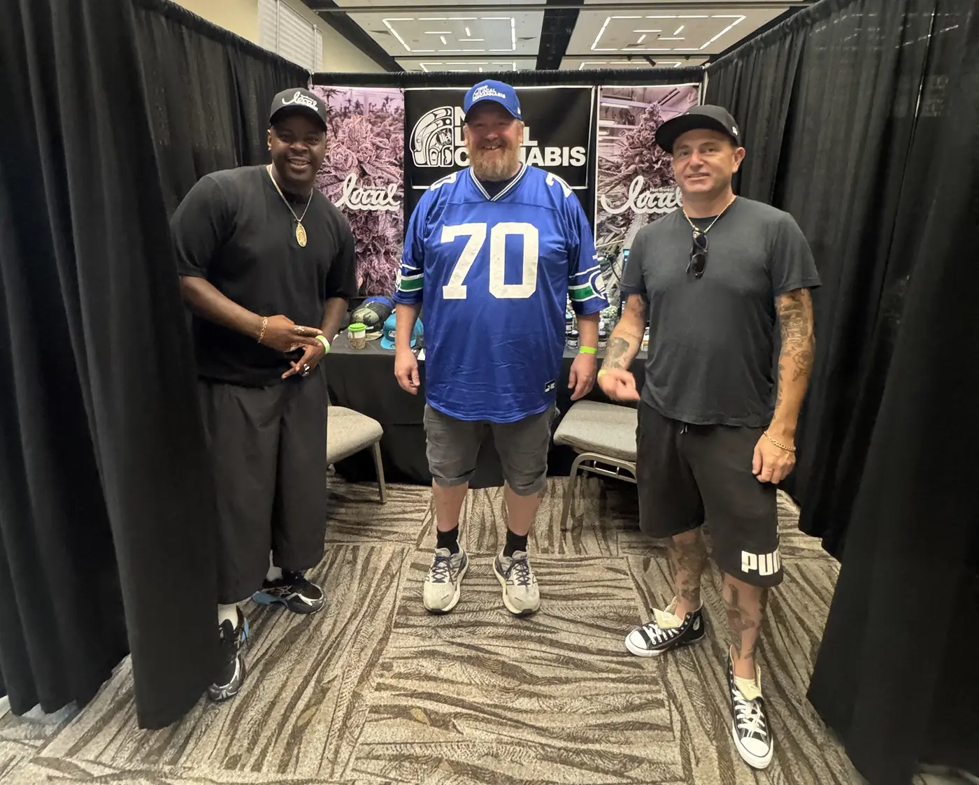 Three people at the booth, one wearing a blue number 70 Seattle jersey, in front of the Northwest Local banners.