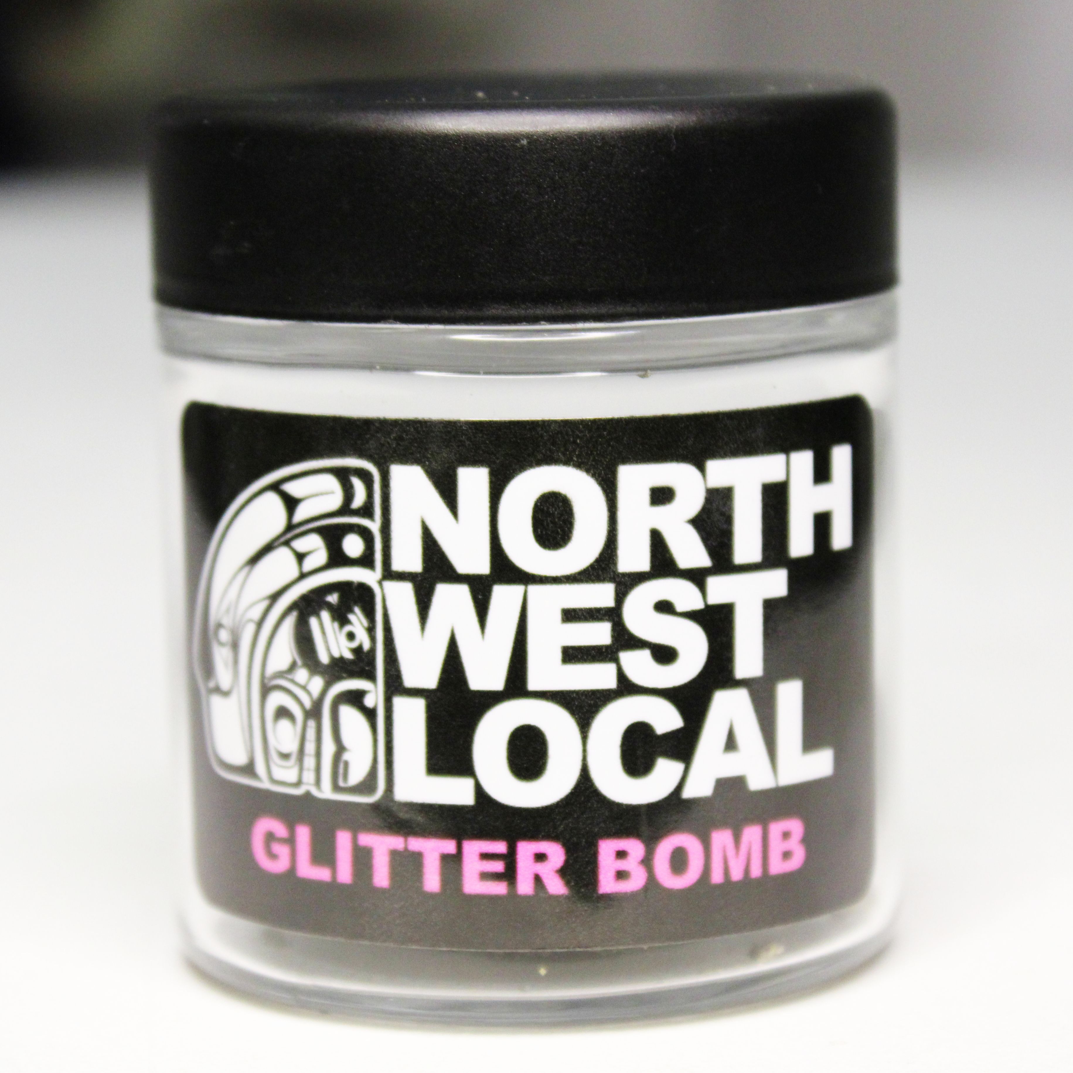 Northwest Local Glitter Bomb eighth in a small glass jar with a black lid, the label showing the formline raven logo and the strain name in pink lettering