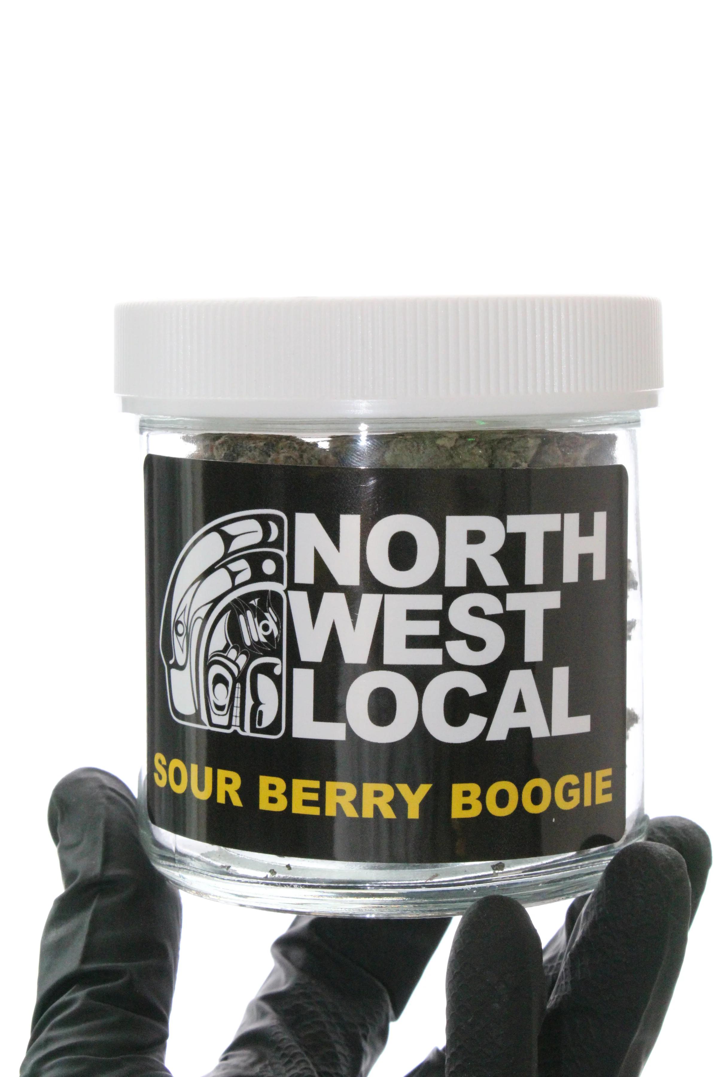 A Sour Berry Boogie jar, its black Northwest Local label facing out over dense flower, held in a gloved hand against white