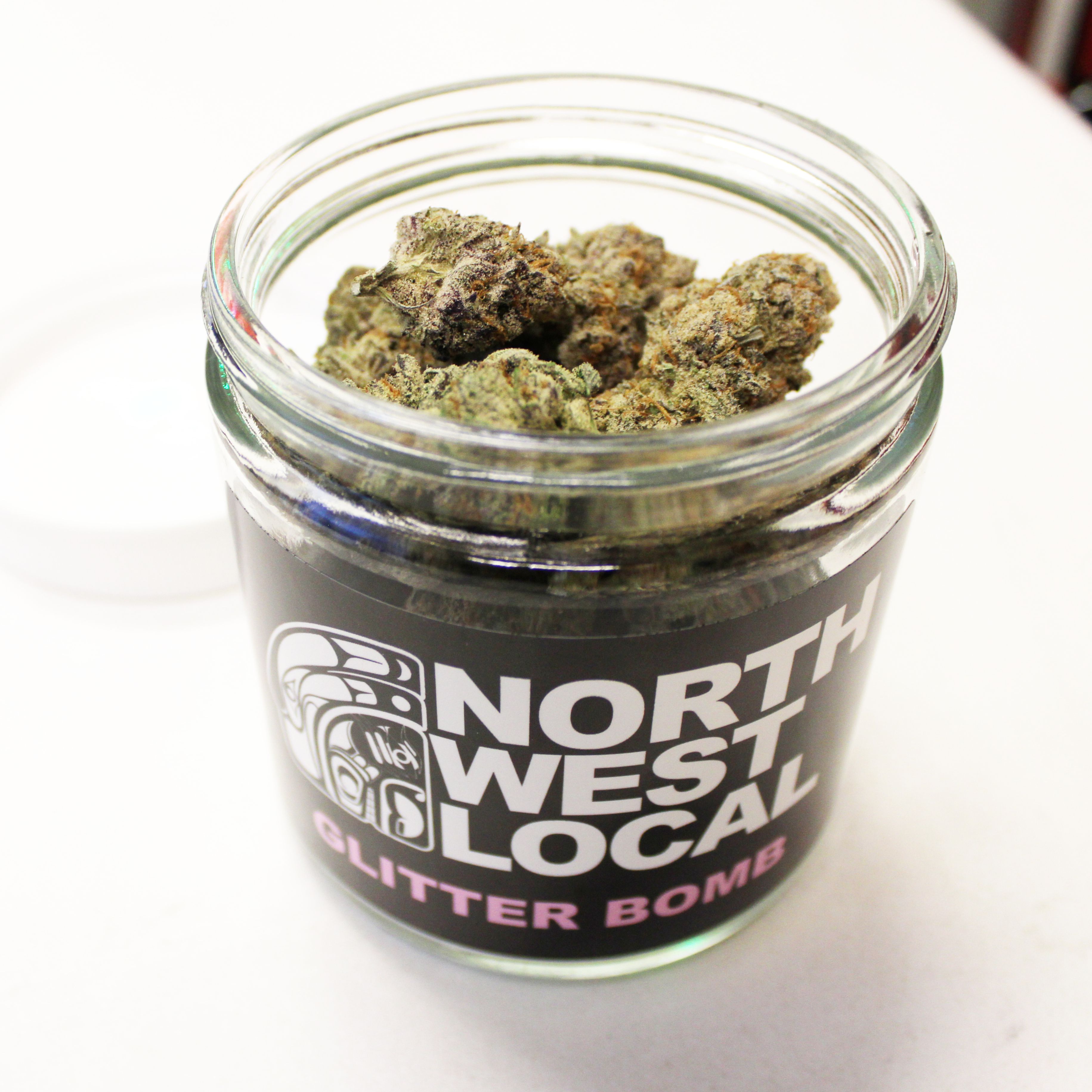 Open Northwest Local Glitter Bomb ounce jar filled with frosty purple and green buds with bright orange pistils, resting above the black label
