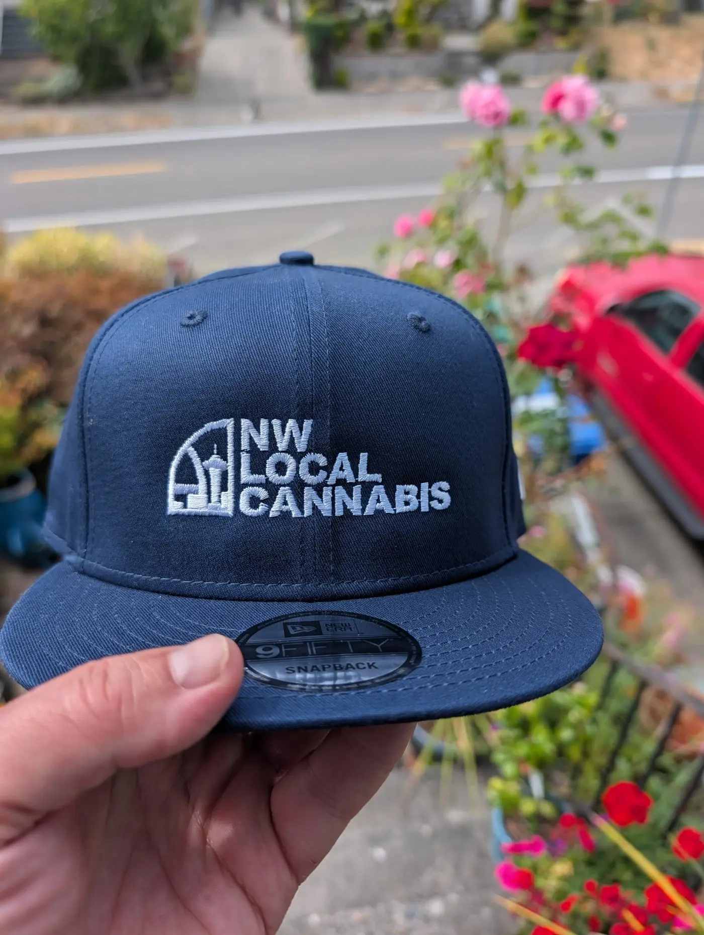 A navy New Era snapback with the NW Local Cannabis block logo, held up outdoors in front of roses.