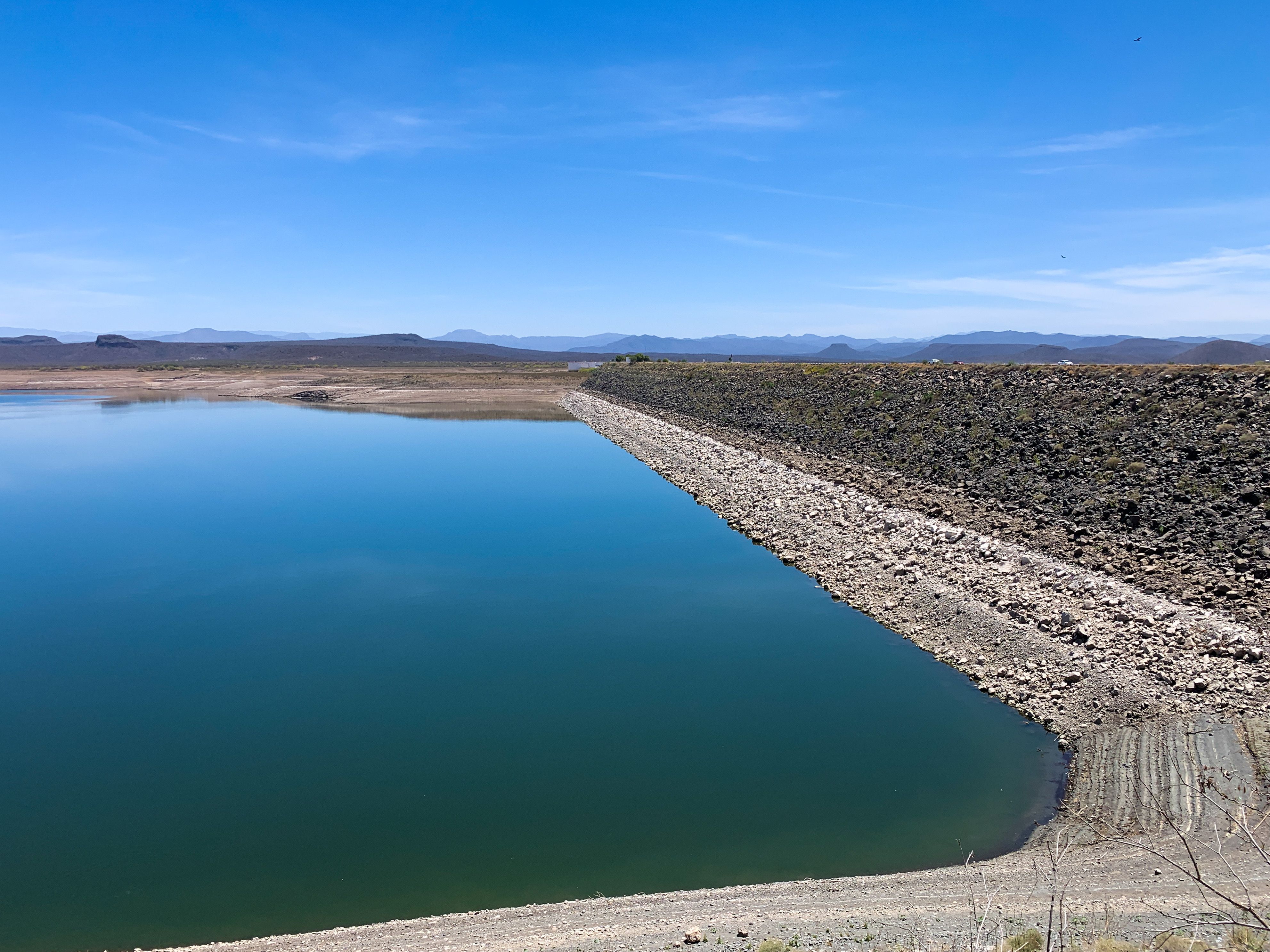 Embalse de la presa Miguel Hidalgo, en Sinaloa: agua azul en calma junto al talud de roca de la cortina, bajo un cielo despejado y con sierras al fondo