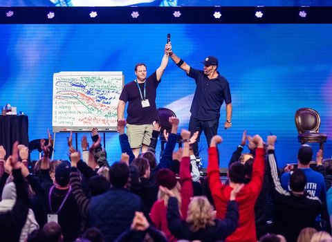 A speaker and another man on stage raise their arms in triumph, mirrored by an audience with raised fists.